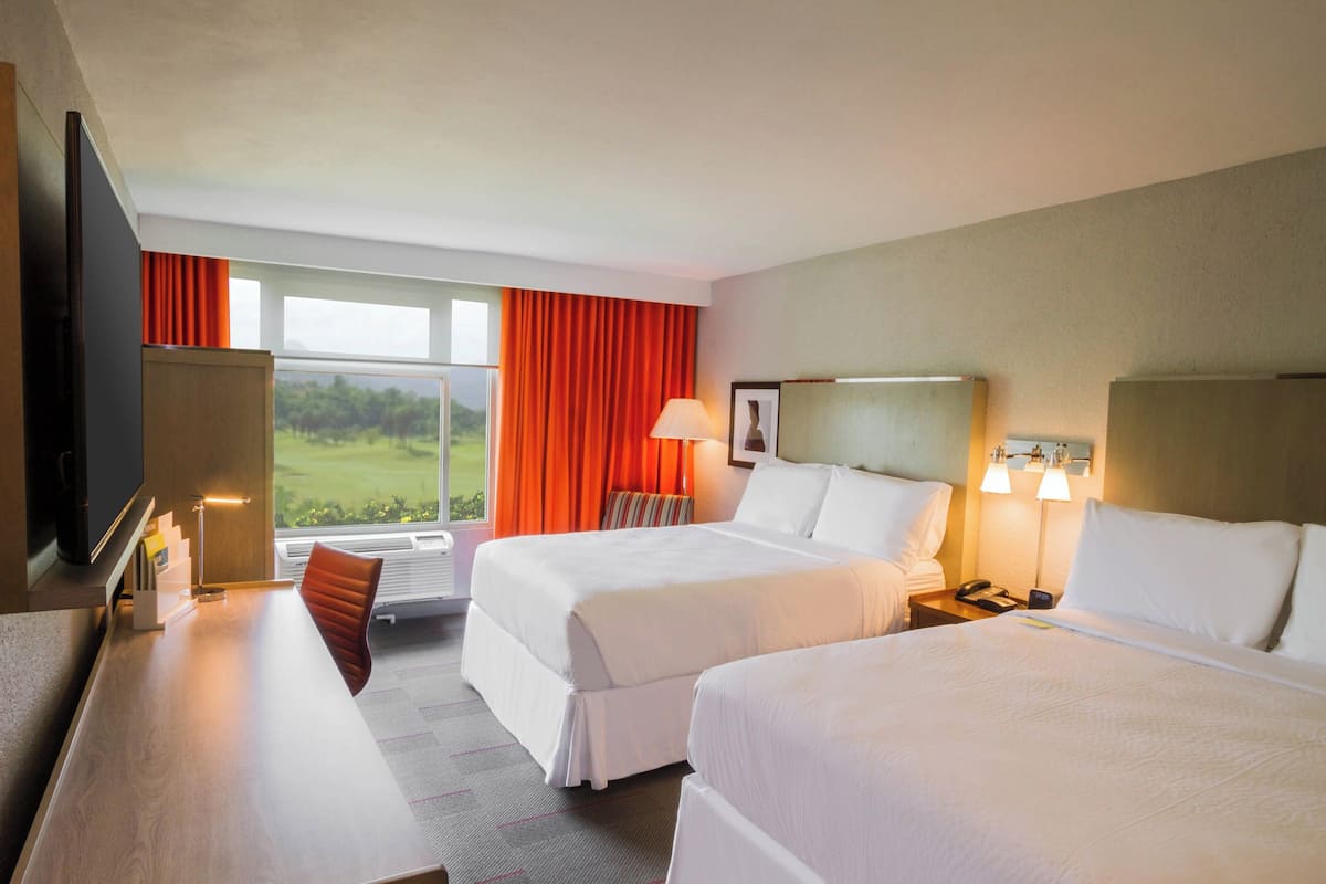 Une chambre d’hôtel avec deux lits, un bureau, une chaise, une télévision, une fenêtre avec des rideaux et une table de chevet avec une lampe.