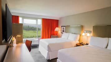 Ein Hotelzimmer mit zwei Betten, einem Schreibtisch, einem Sessel, einem Fernseher, einem Fenster mit Vorhängen und einem Nachttisch mit Lampe.