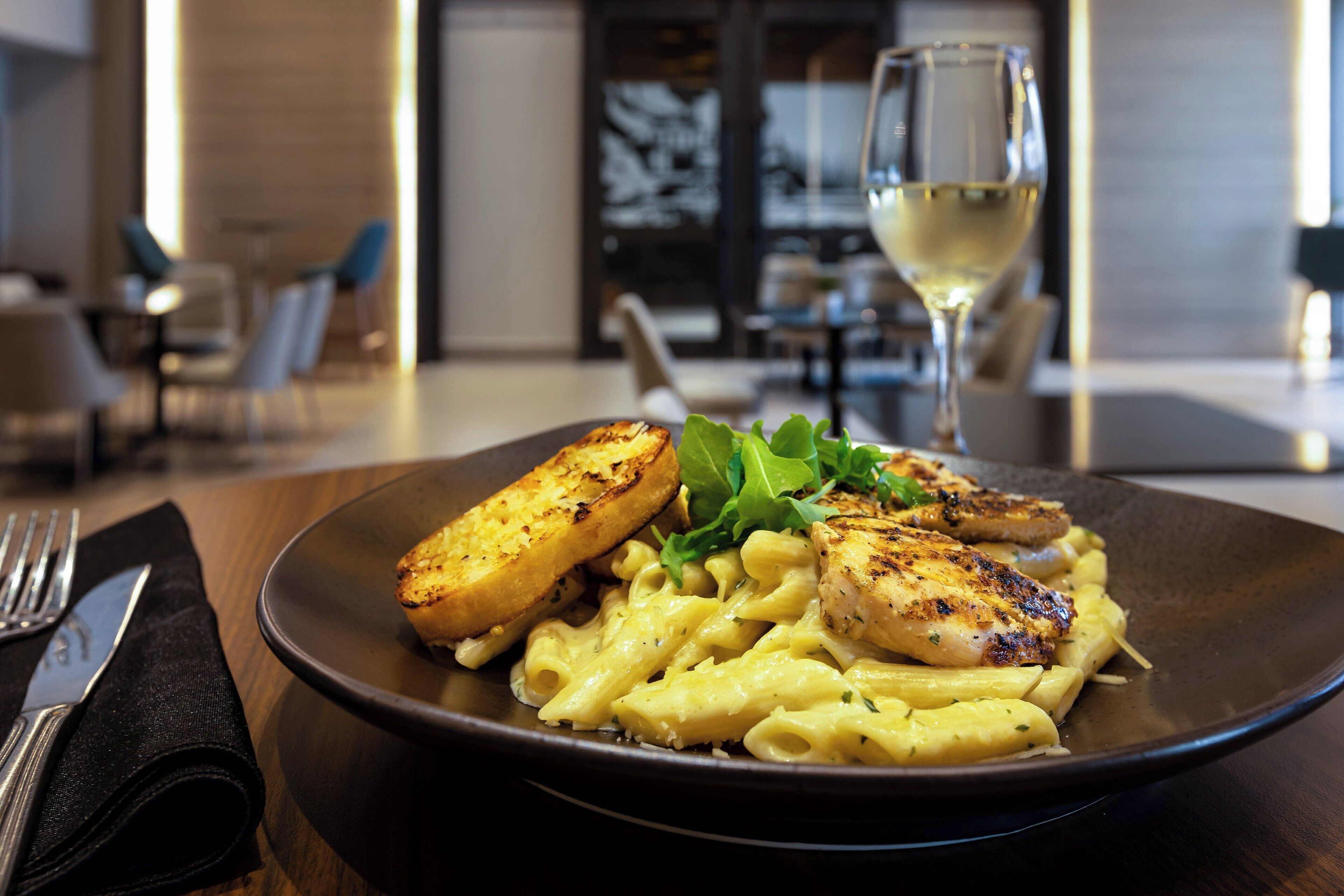 En tallrik pasta med grillad kyckling och rostat bröd, serverad tillsammans med ett glas vitt vin, på ett träbord i en modern matsal.
