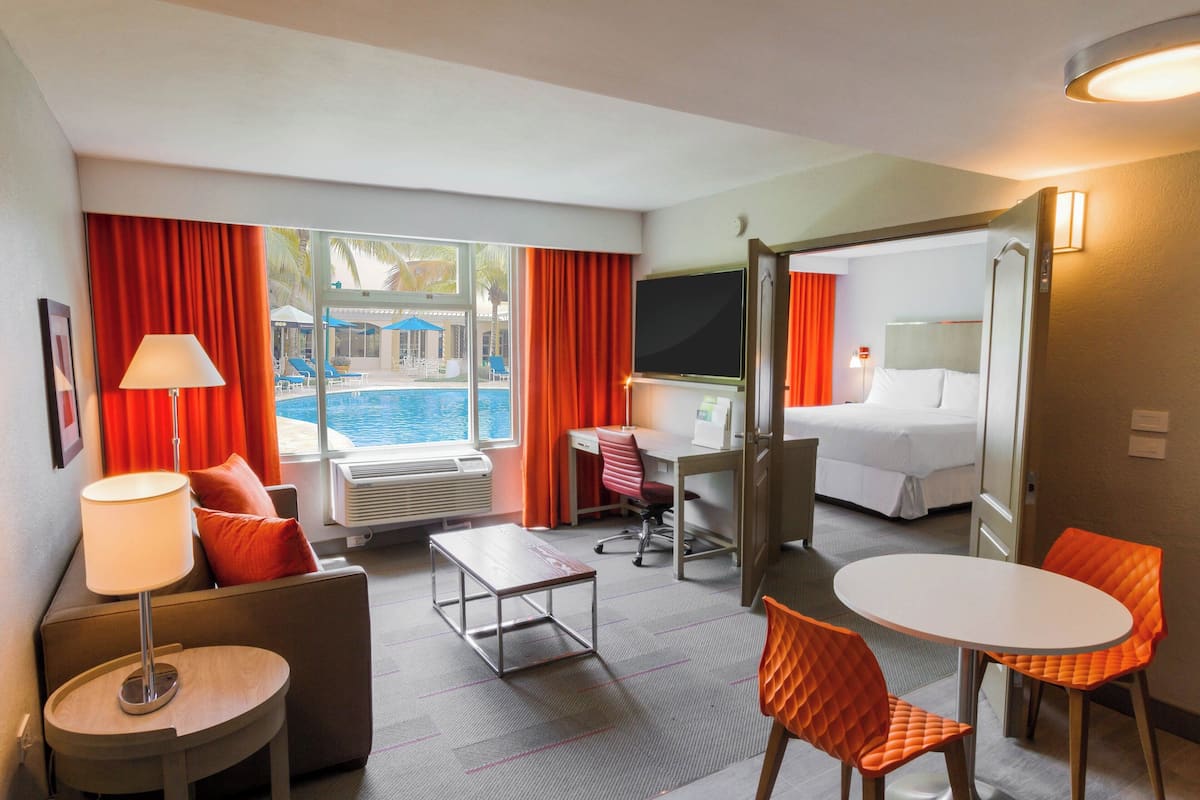 Une chambre d’hôtel avec un lit, un bureau et un coin salon. Une fenêtre offre une vue sur une piscine et des palmiers.
