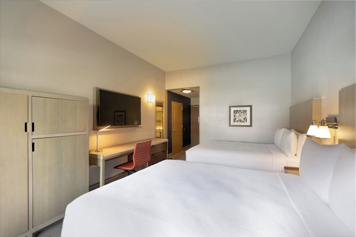 Une chambre d’hôtel avec deux lits, un bureau, une chaise, une armoire et un tableau encadré au mur.