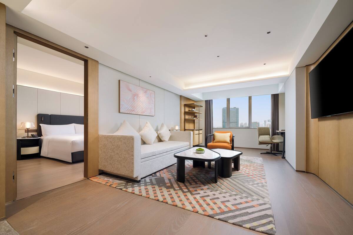 A modern hotel room with a bed, sofa, coffee table, TV, and city view.