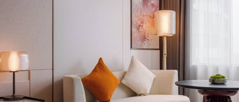 Habitación de hotel moderna con sofá, dos lámparas, una obra de arte enmarcada, una mesita con una planta y una ventana con cortinas.