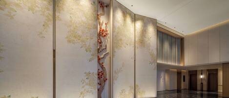 A spacious lobby with a polished marble floor, a large decorative tree with red flowers, and a modern elevator.