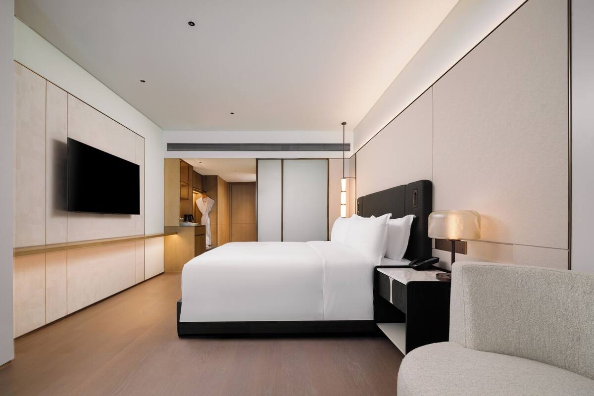 A modern hotel room with a large bed, a TV mounted on the wall, a bedside table with a lamp, and a chair.