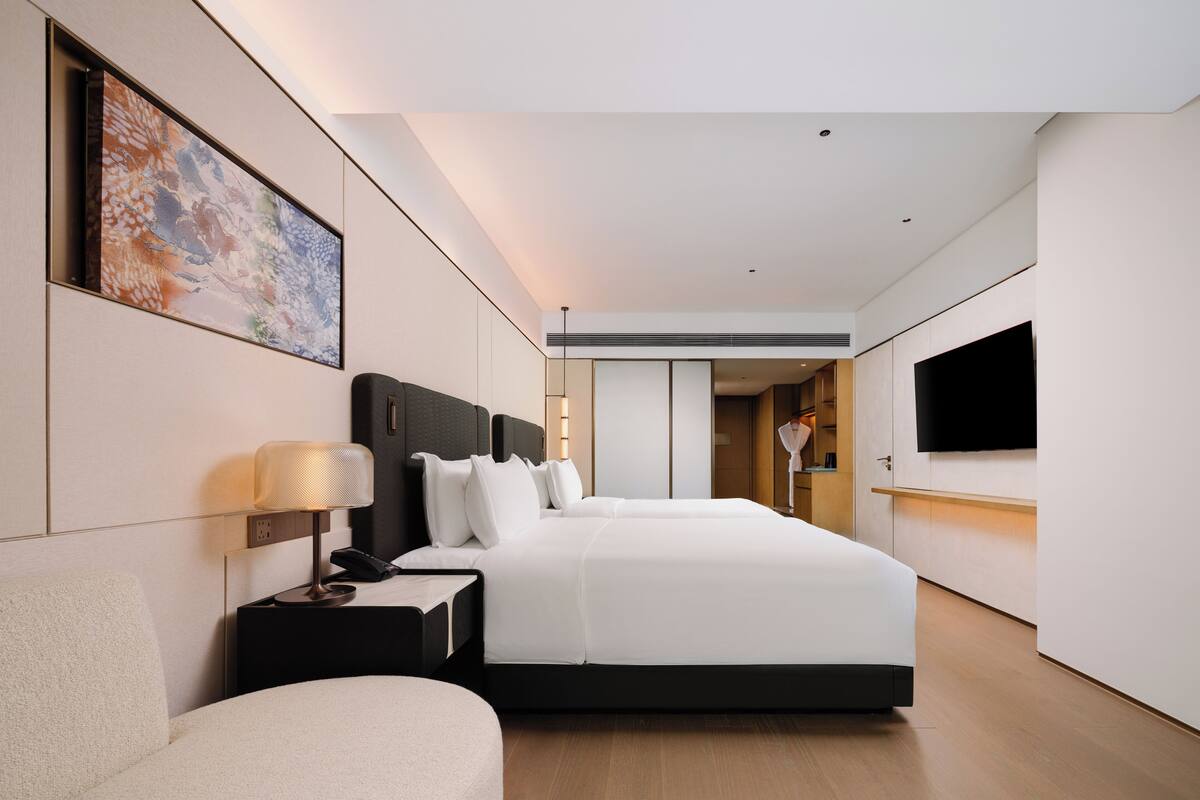 A modern hotel room with a large bed, bedside table, lamp, and a flat-screen TV mounted on the wall.