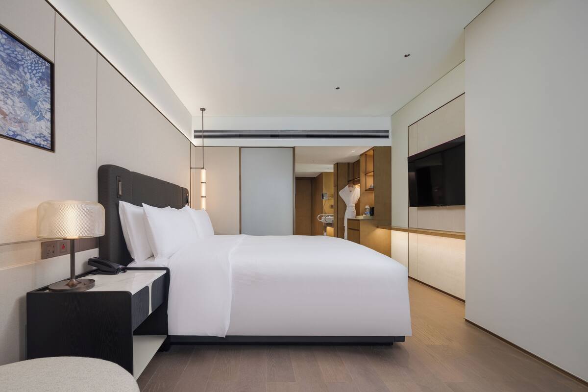 A modern hotel room with a large bed, bedside table, lamp, and a TV mounted on the wall.
