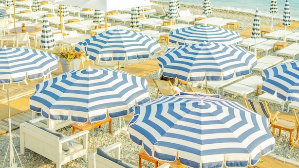 A beach with rows of white lounge chairs and blue-striped umbrellas, some with white canopies, arranged on a pebbled surface. - Hotel Aria (Nice)