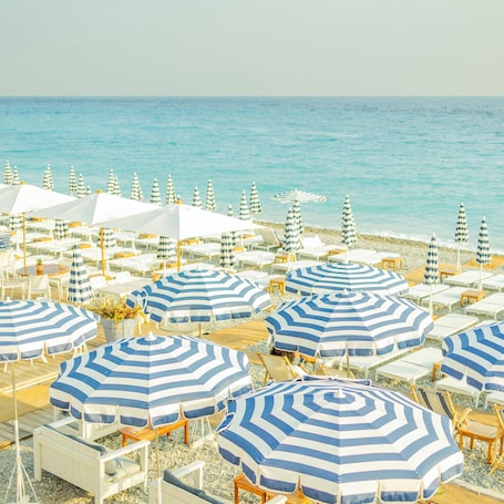 A beach with rows of white lounge chairs and blue-striped umbrellas, some with white canopies, arranged on a pebbled surface.