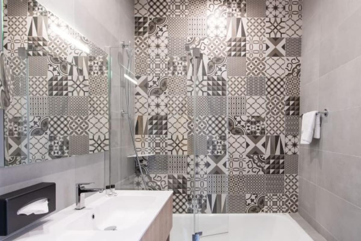 A modern bathroom with a large mirror, a white sink, and a shower with a geometric tile pattern.