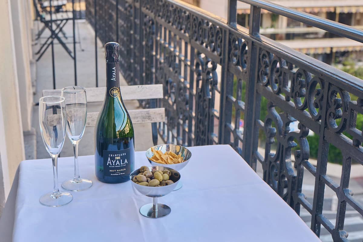 A balcony table with a bottle of champagne, two glasses, a bowl of olives, and a white tablecloth.