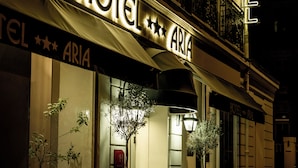Un hotel llamado "HOTEL ARIA" con un letrero iluminado llamativo, un balcón con barandillas de metal ornamentadas y una planta en maceta frente a la entrada.
