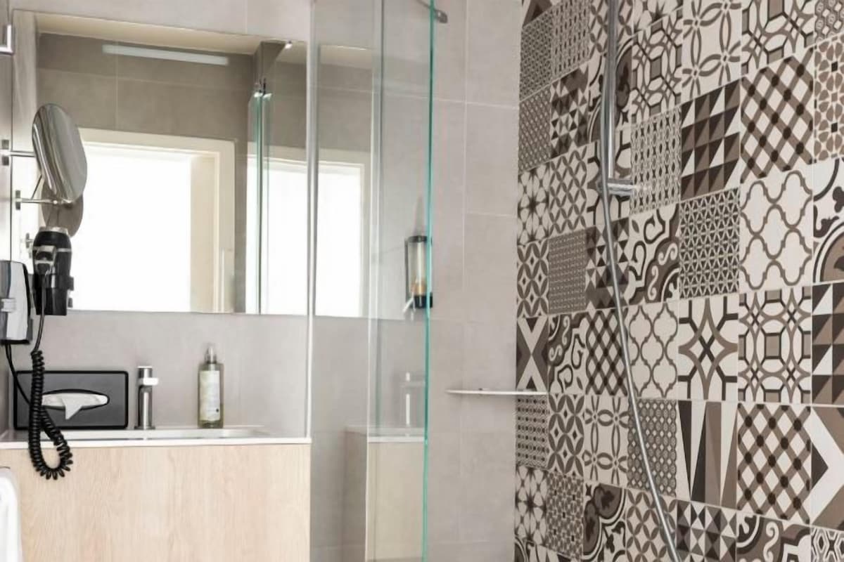 Un baño con cabina de ducha de vidrio, un lavabo con espejo y un revestimiento de pared de azulejos estampados.