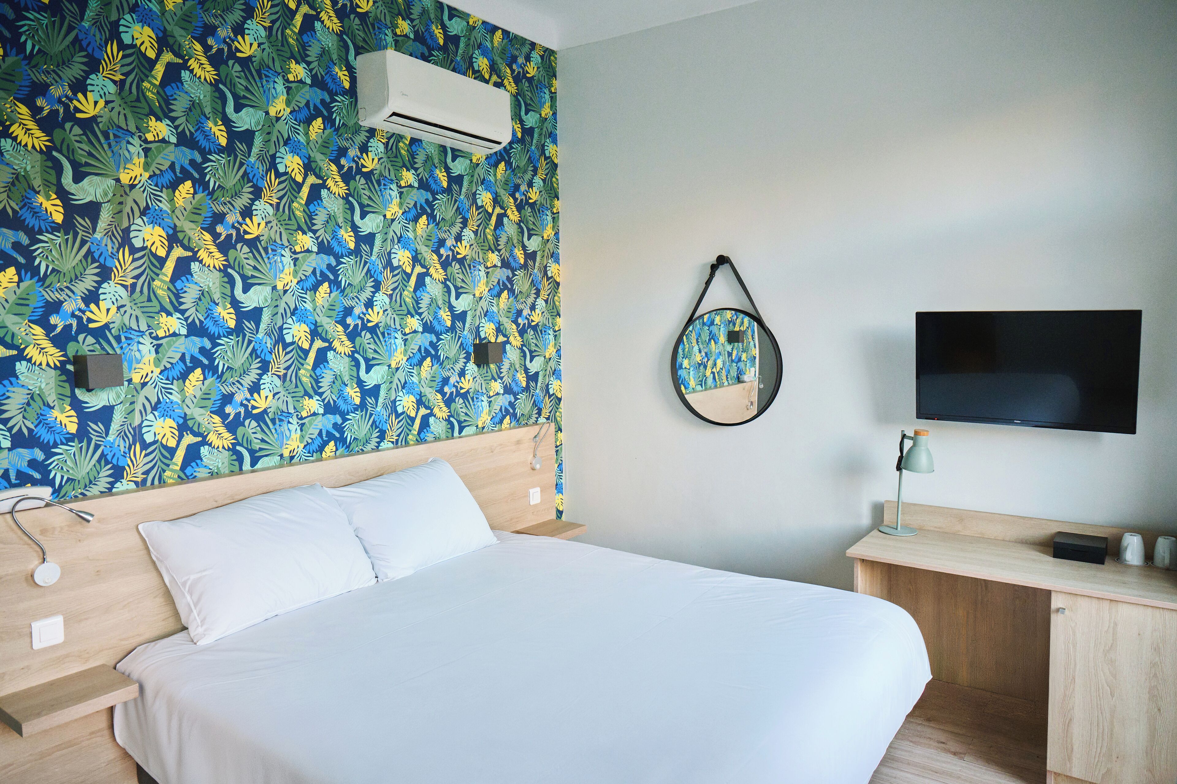 A hotel room with a bed, a desk, a TV, an air conditioner, and a wall with a tropical leaf pattern.