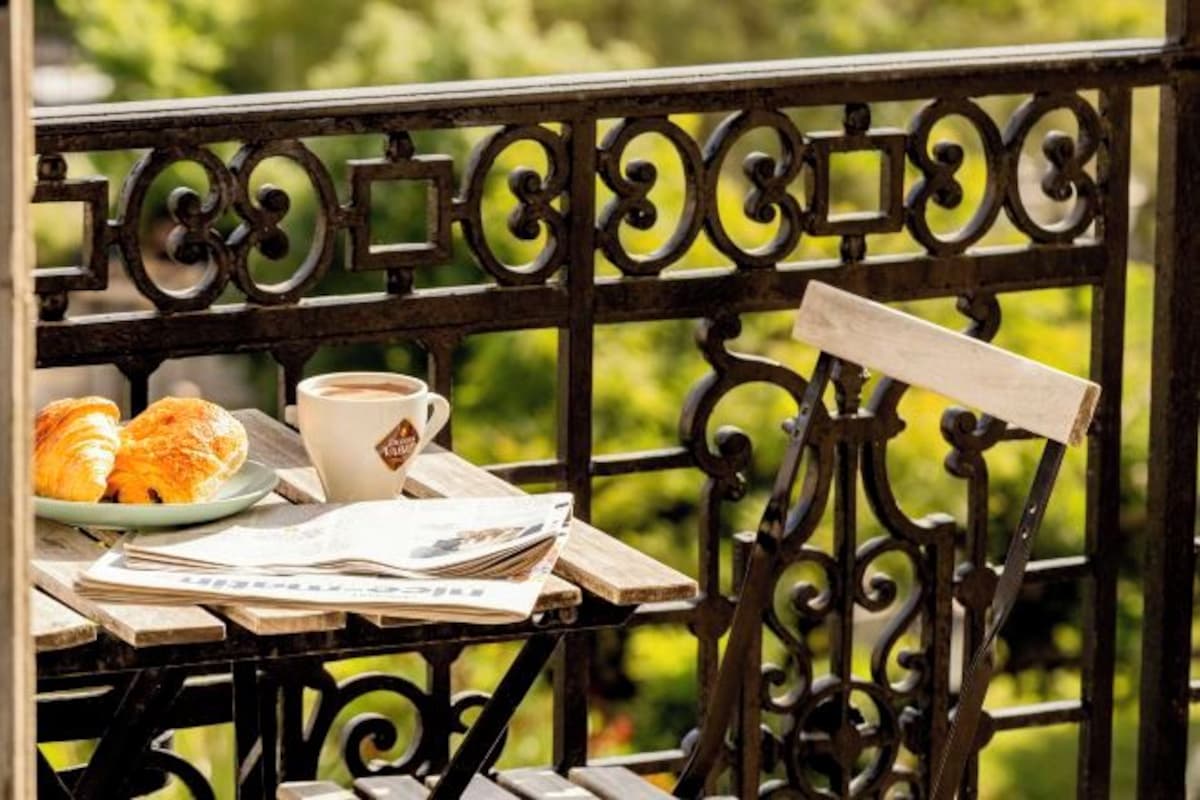 A table with croissants, a cup of coffee, and newspapers on a balcony with ornate metal railings.