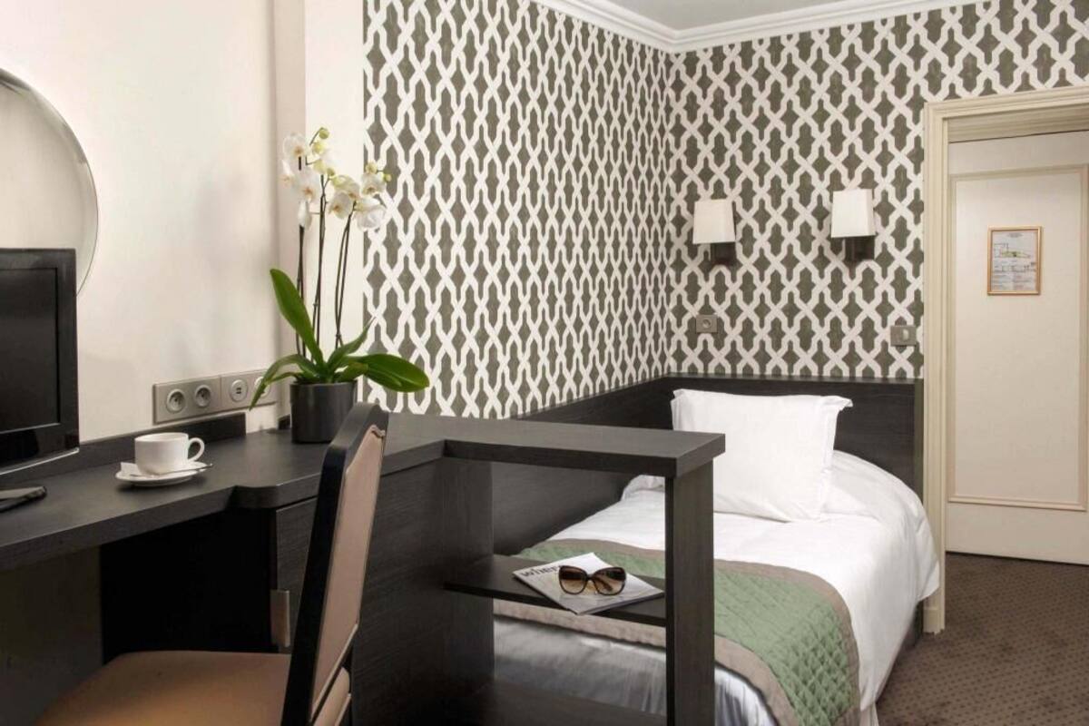 A hotel room with a bed, a desk with a cup and a plant, a chair, a TV, and a patterned wall.