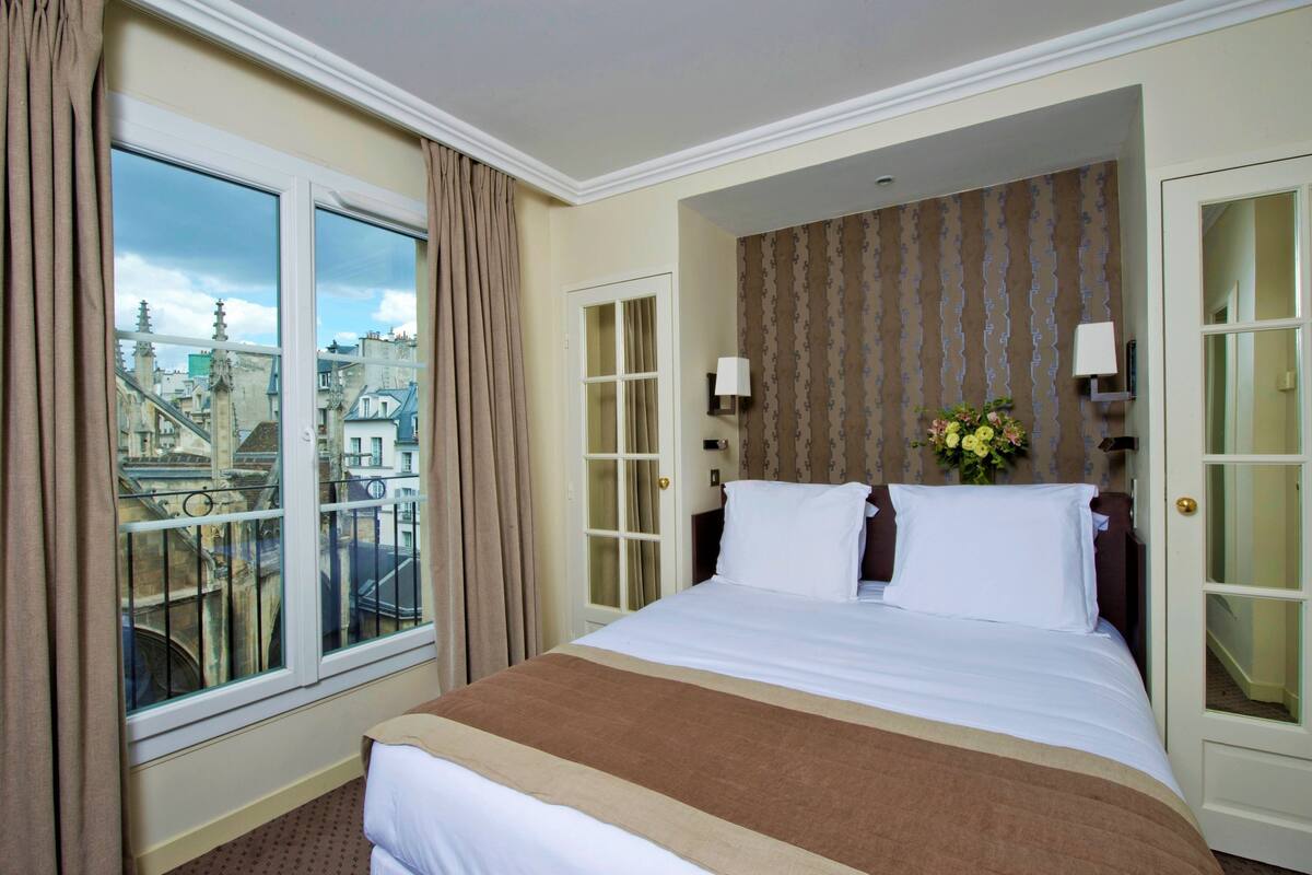 A hotel room with a large bed, a window with curtains, a view of buildings, and a patterned wallpaper.