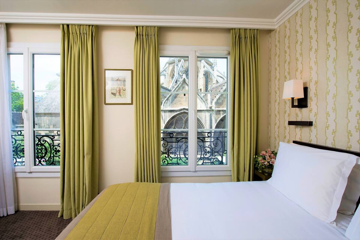 A hotel room with a bed, green curtains, a window with a view of a building, and a wall-mounted lamp.