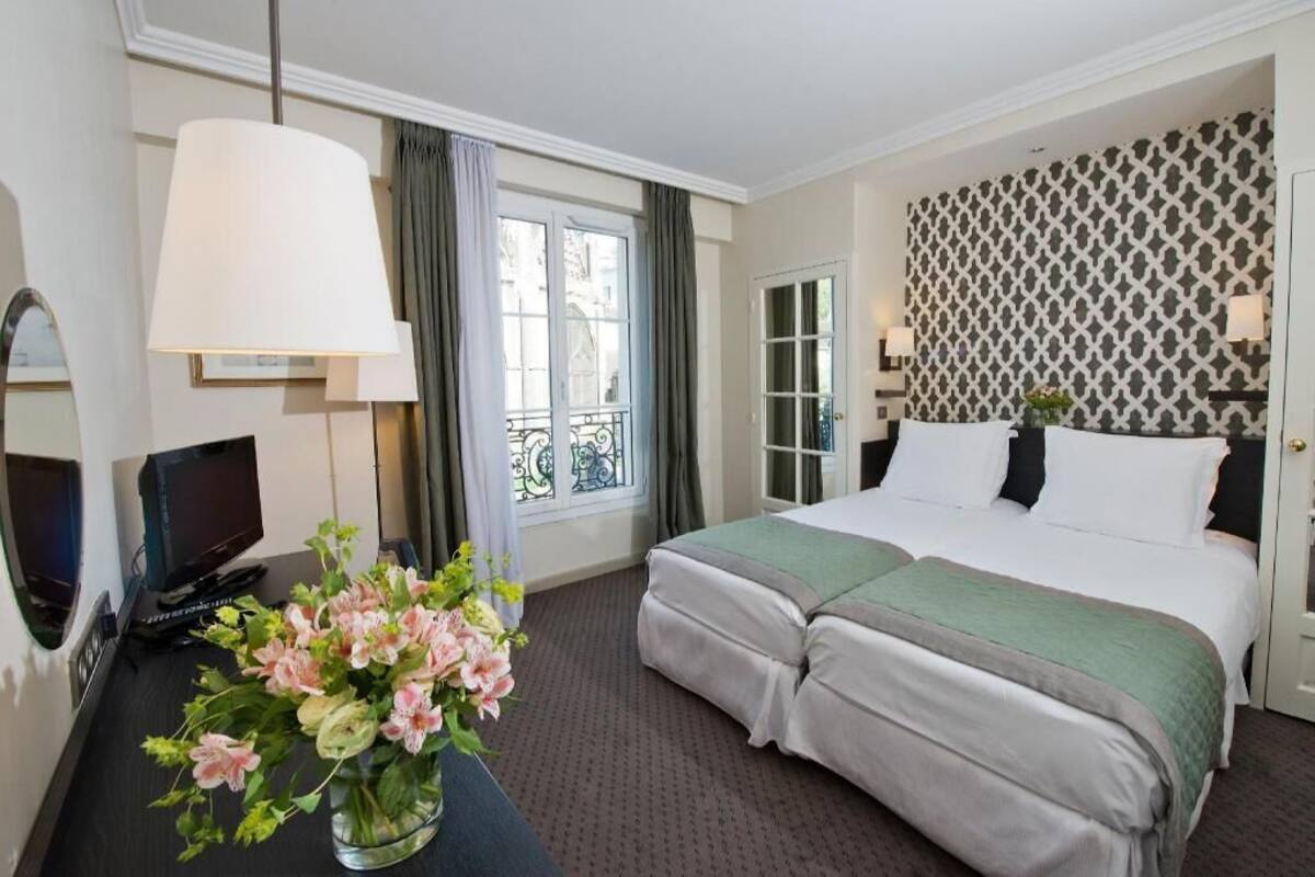 Habitación de hotel con cama, televisión, escritorio con un jarrón de flores, ventana con cortinas y pared con diseño.