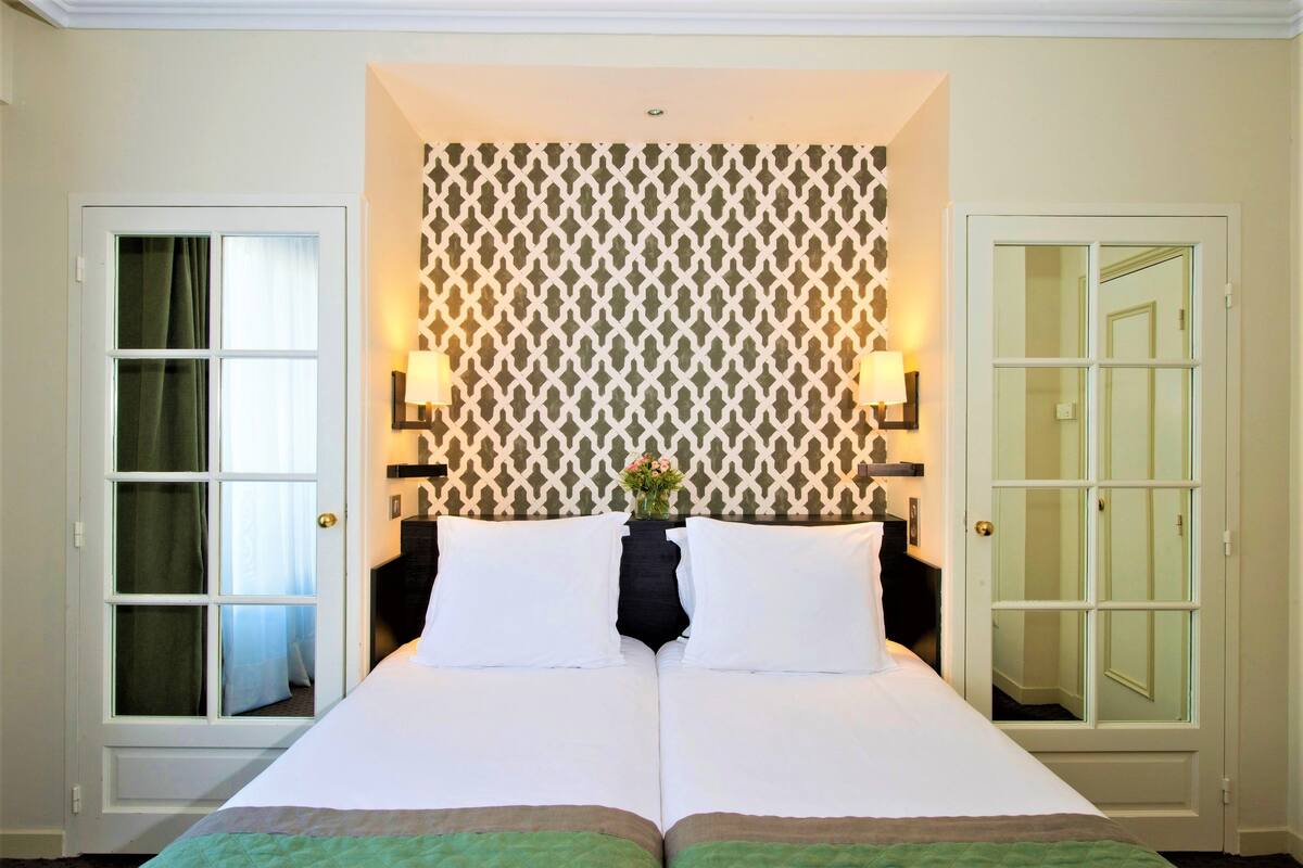 A double bed with white linens, two bedside tables with lamps, a patterned wall, and a door with a mirror.
