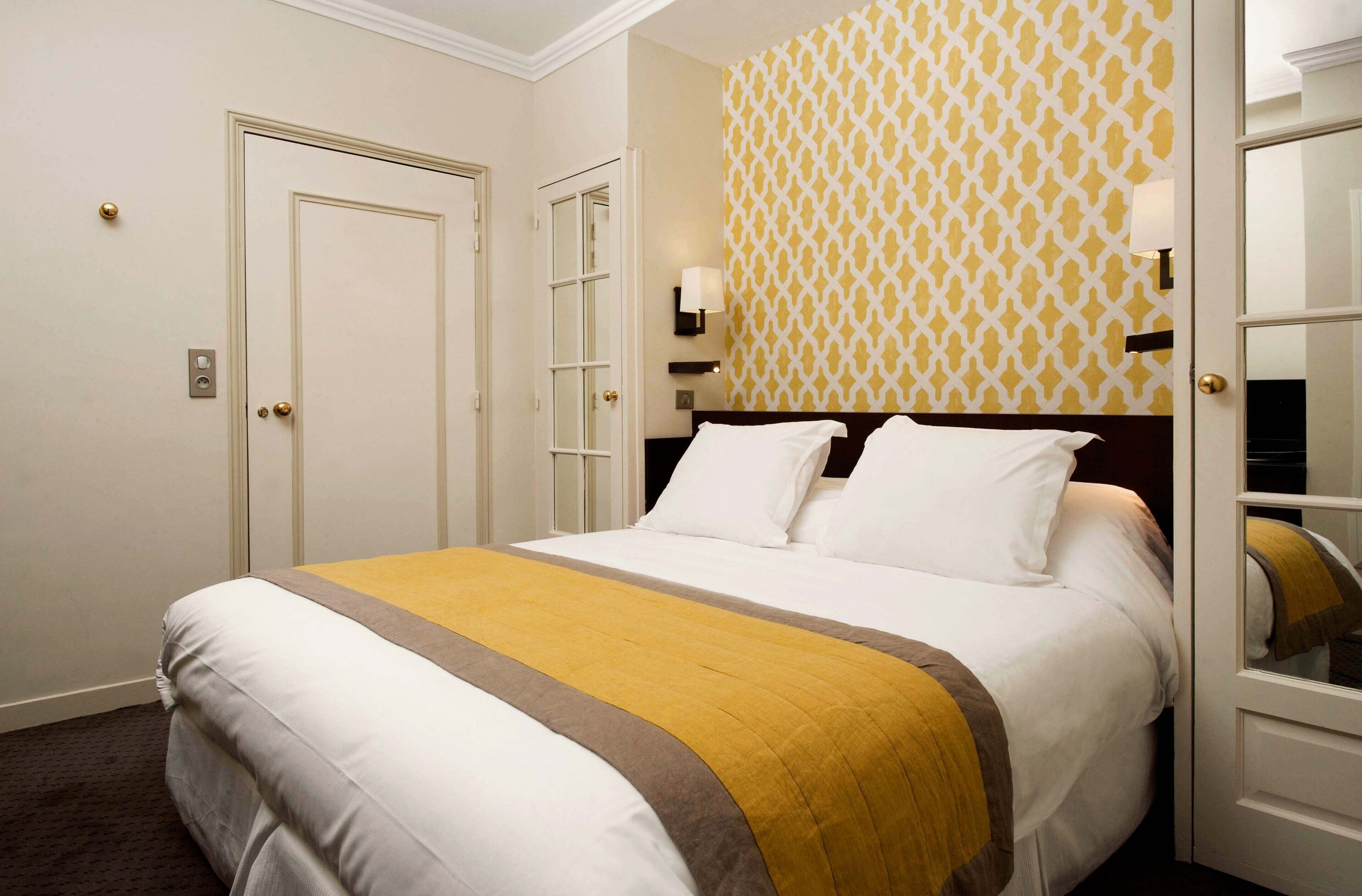Un lit bien fait, recouvert d’une couvre-lit blanche et jaune, avec une tête de lit en bois et un mur à motifs derrière.