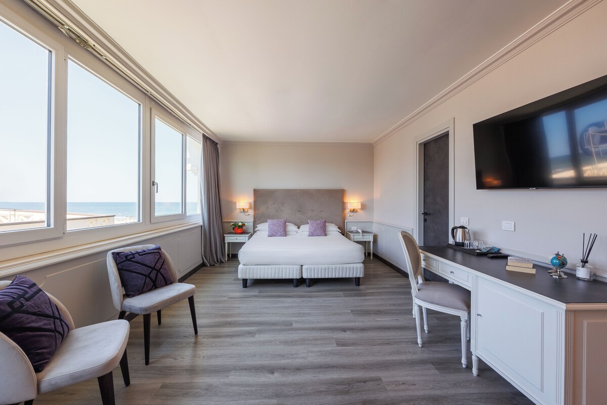 A hotel room with a large bed, a desk, a chair, a TV, and a view of the beach.