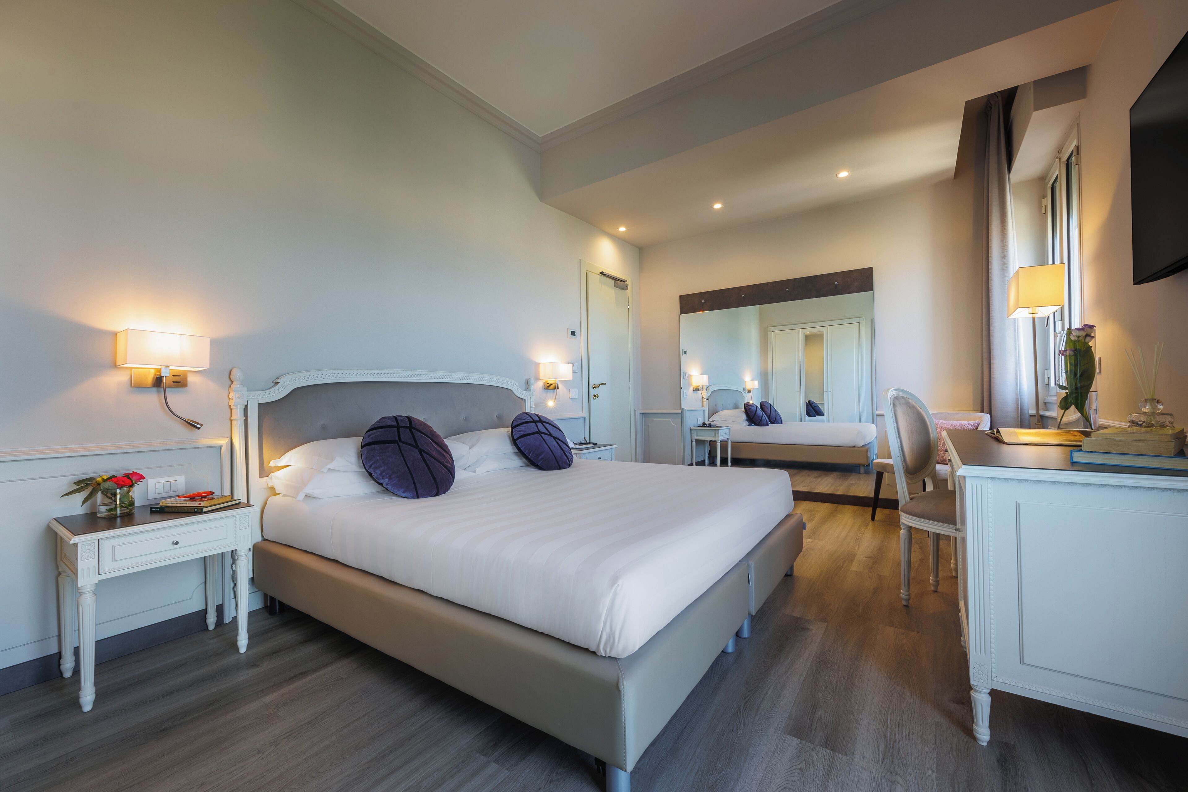 A modern hotel room with a large bed, bedside tables, a desk, and a mirror.