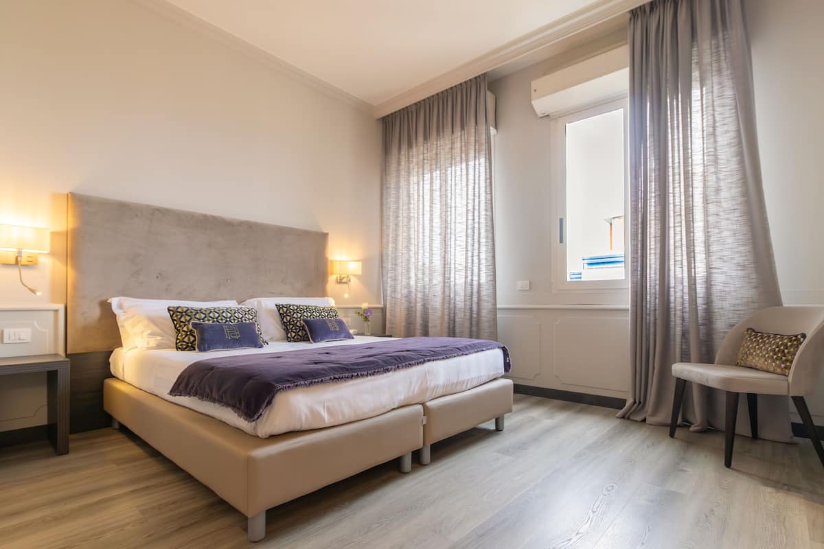 A modern bedroom with a large bed, bedside lamps, a chair, and a window with curtains.