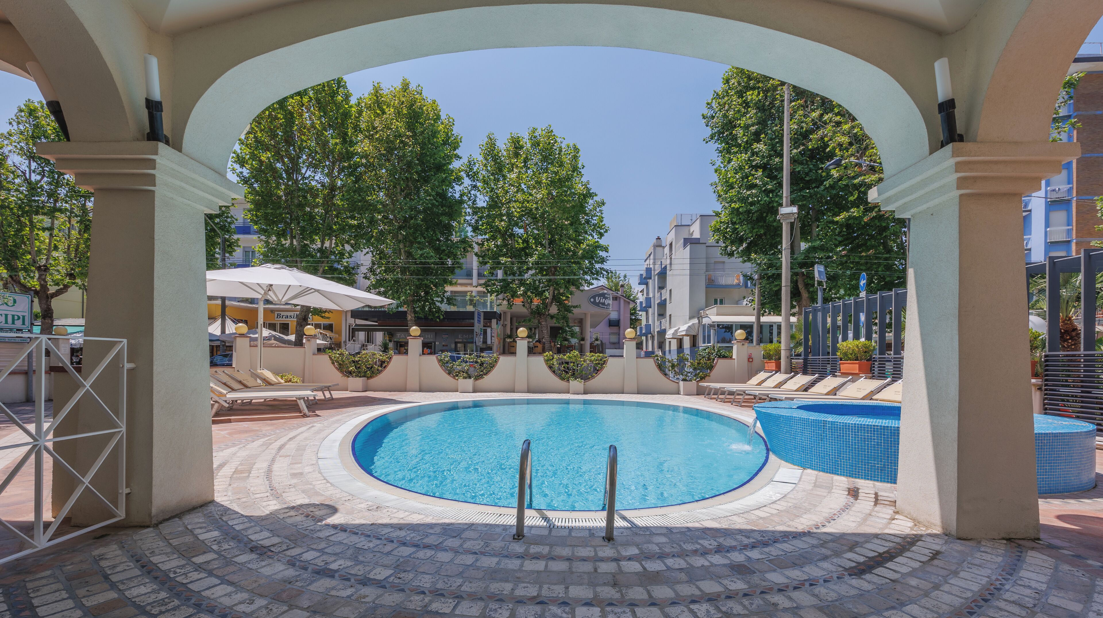 Une piscine dotée d’une terrasse carrelée, entourée de chaises longues et de parasols, et située sous une zone abritée soutenue par des colonnes.