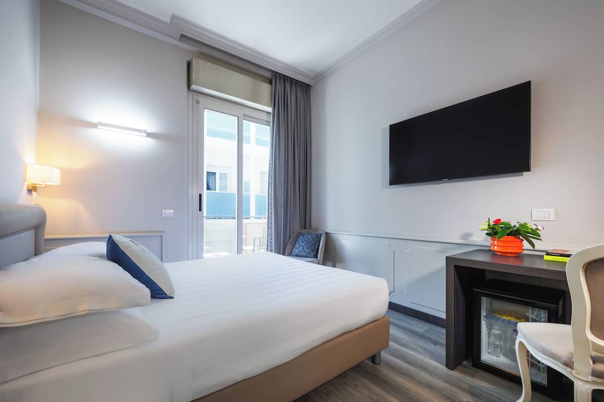 A hotel room with a bed, a desk, a chair, a flat-screen TV, a window with curtains, and a view of the outside.