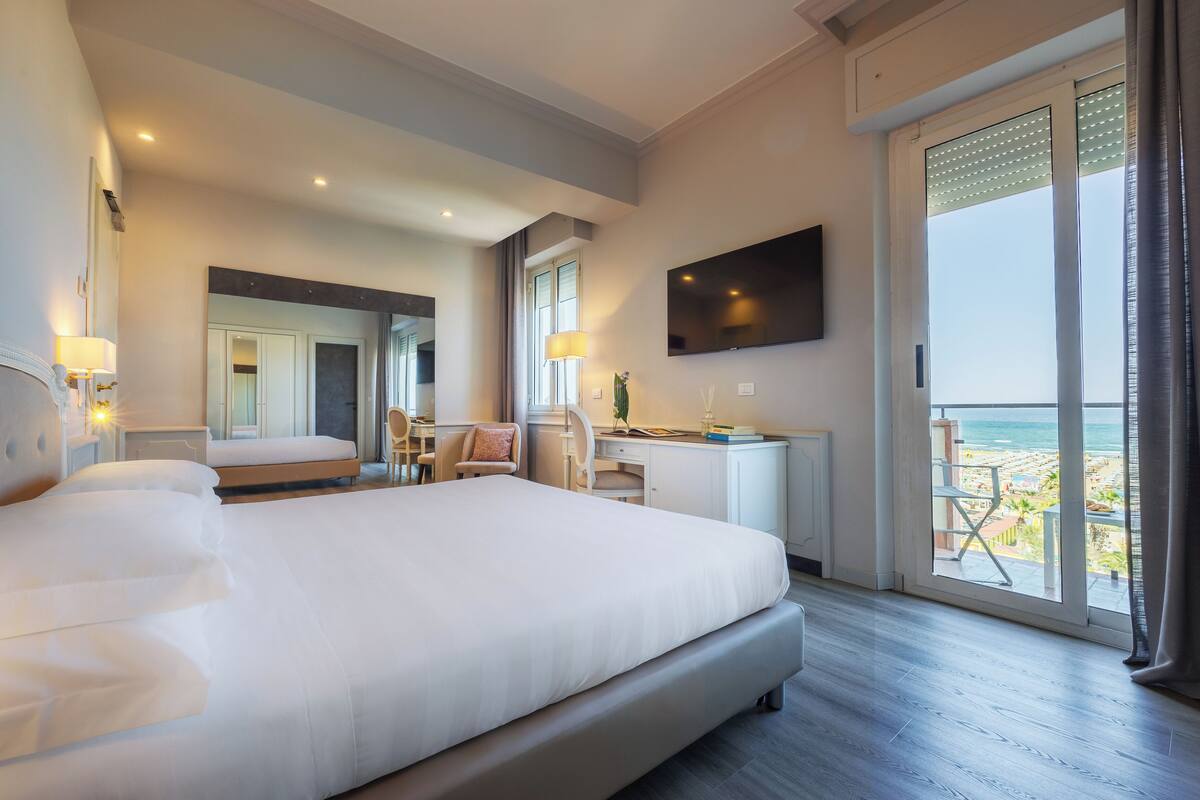 A hotel room with a large bed, a desk, a chair, a TV, and a balcony with a view of the beach.