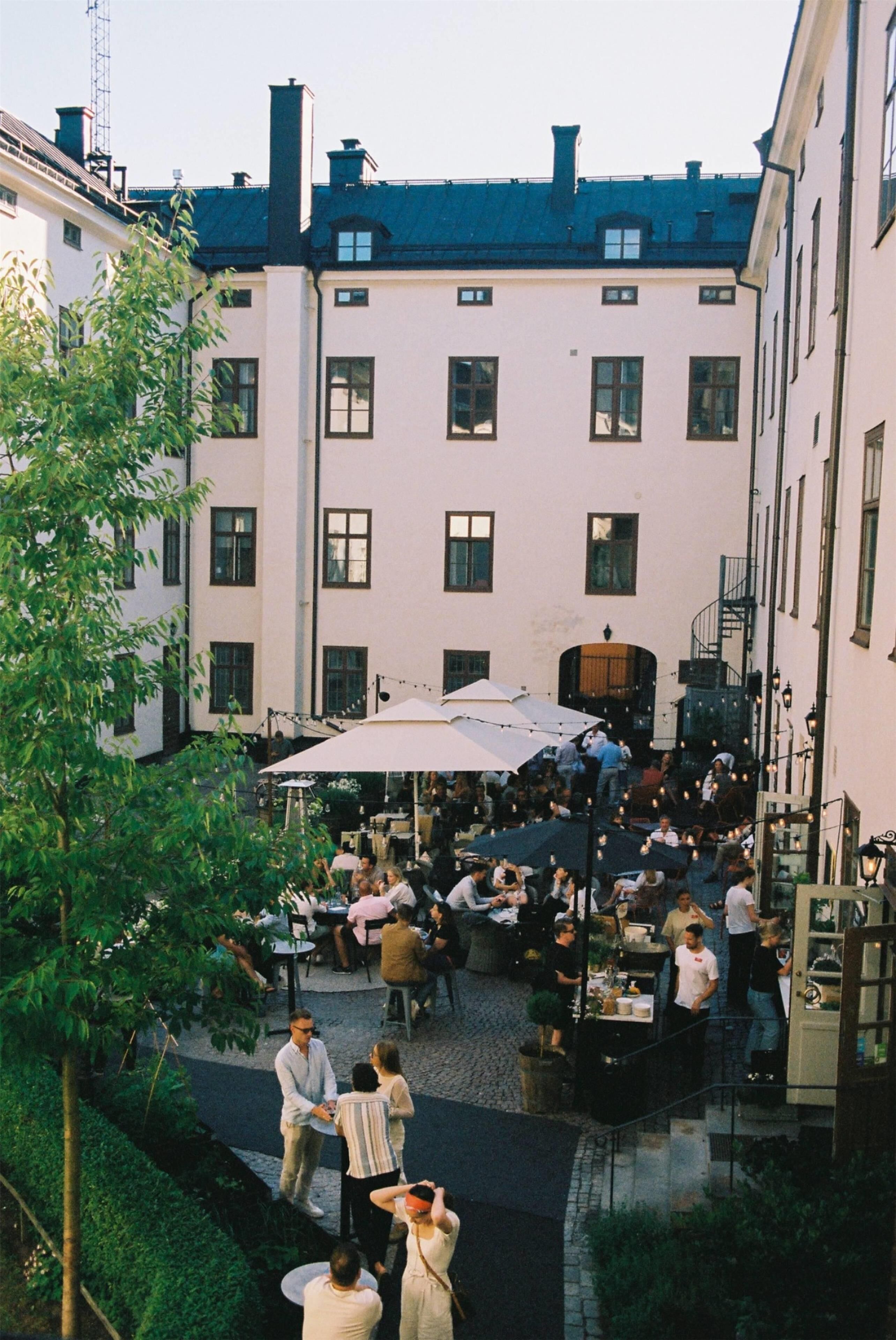 Ein Innenhof mit Außenbestuhlung, umgeben von Gebäuden mit zahlreichen Fenstern und einer Treppe, die zu einer oberen Ebene führt.