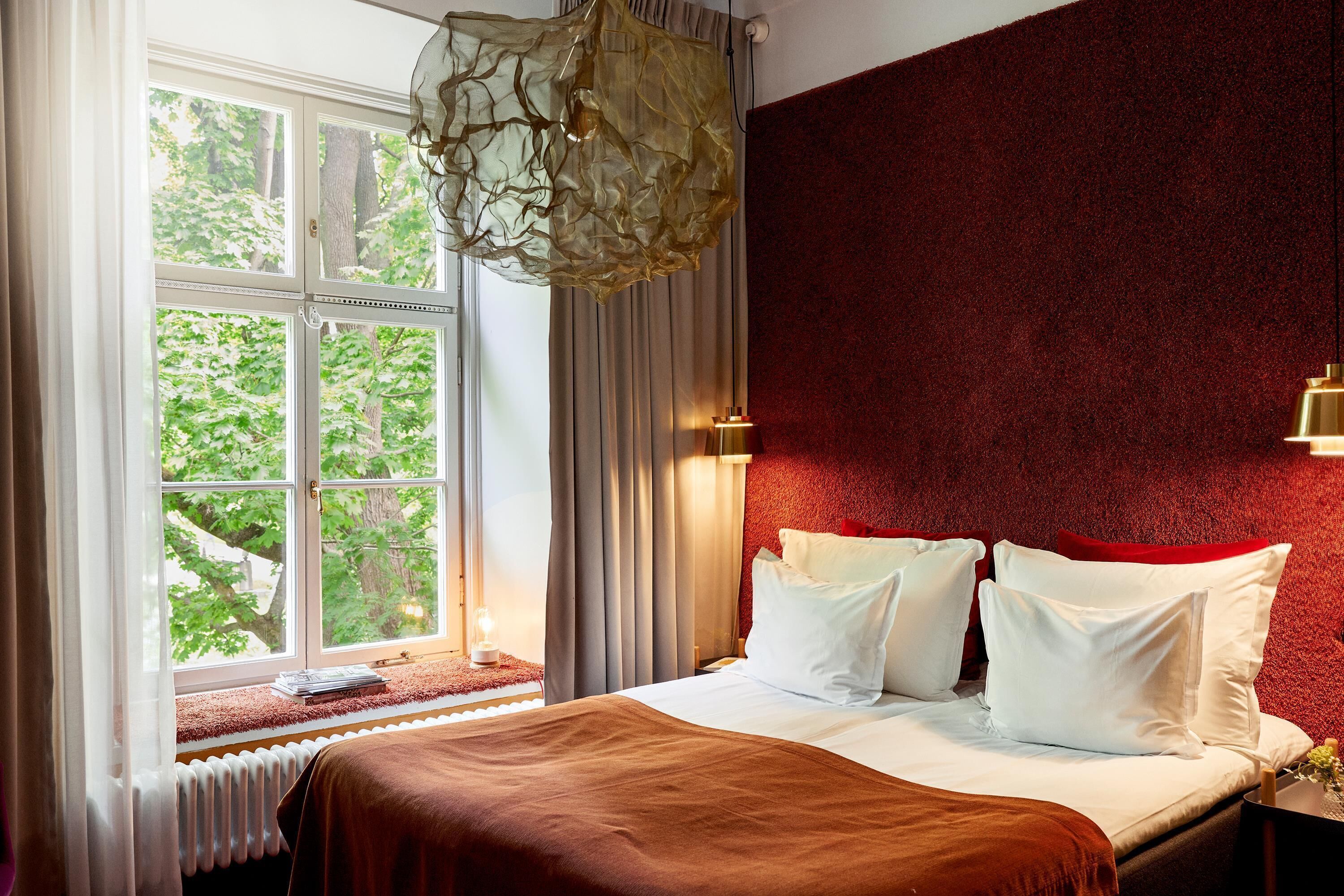 A hotel room with a bed, a window with curtains, a radiator, and a red textured wall.