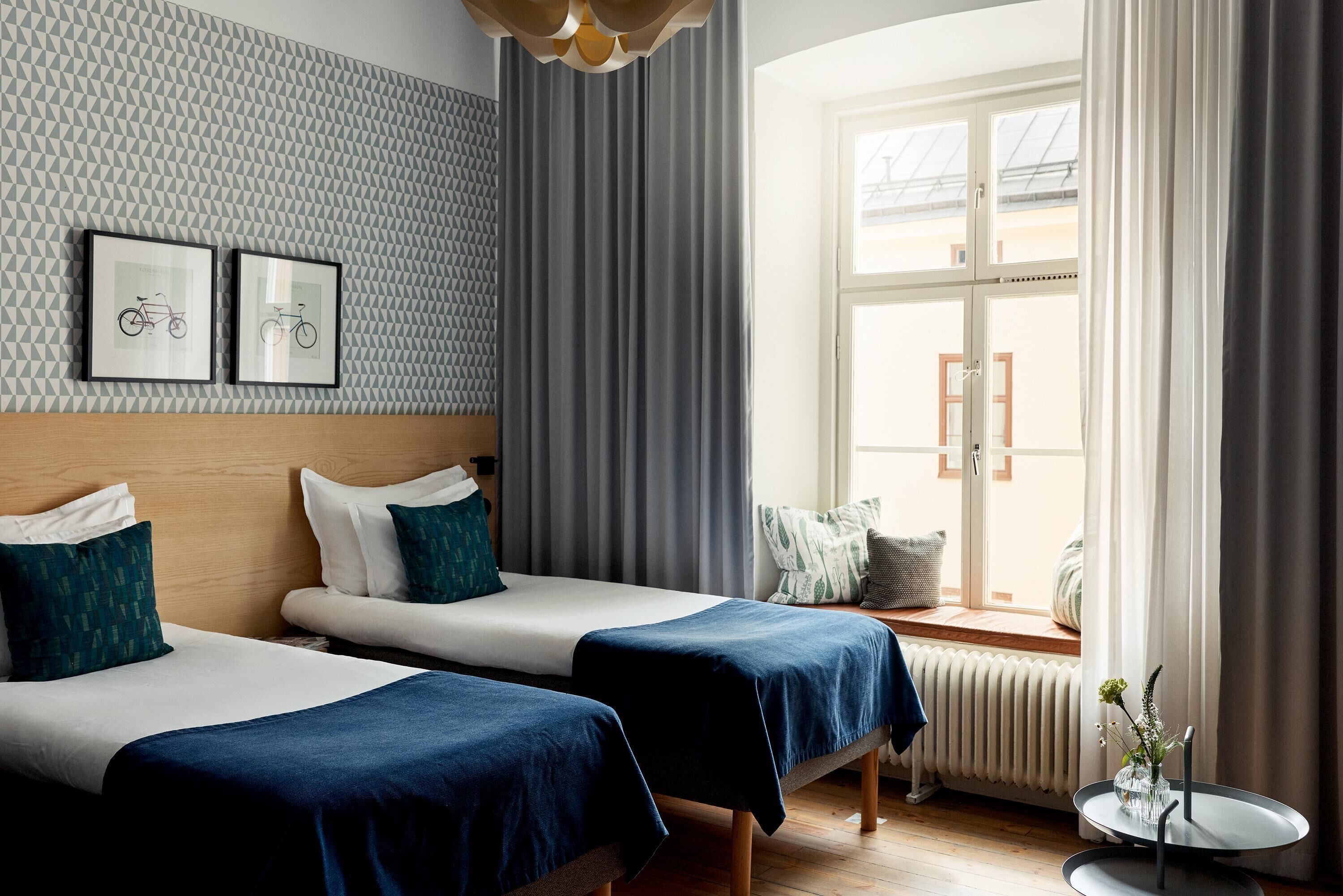 A hotel room with two beds, a window with curtains, a radiator, and two framed pictures on the wall.