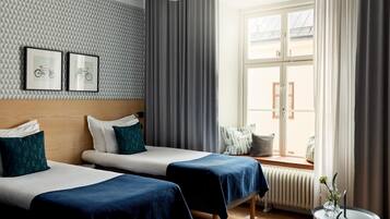 A hotel room with two beds, a window with curtains, a radiator, and two framed pictures on the wall.