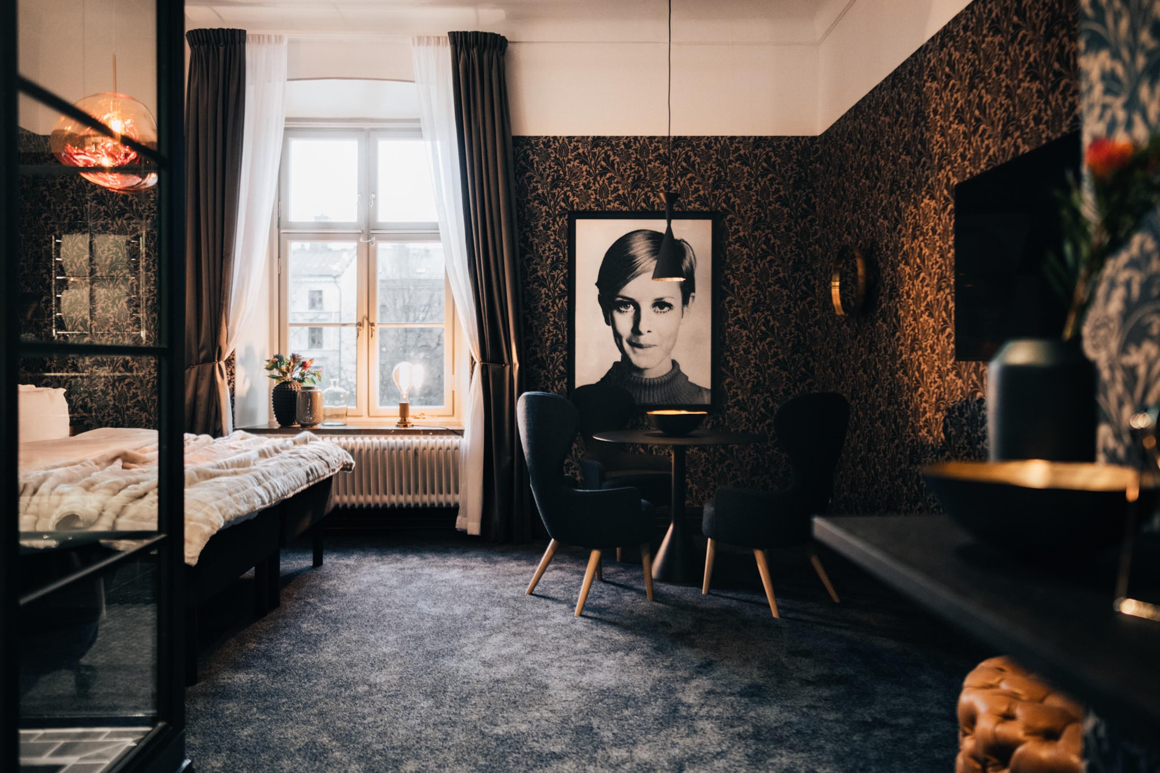 A hotel room with a bed, a desk, a chair, a radiator, and a window with curtains.