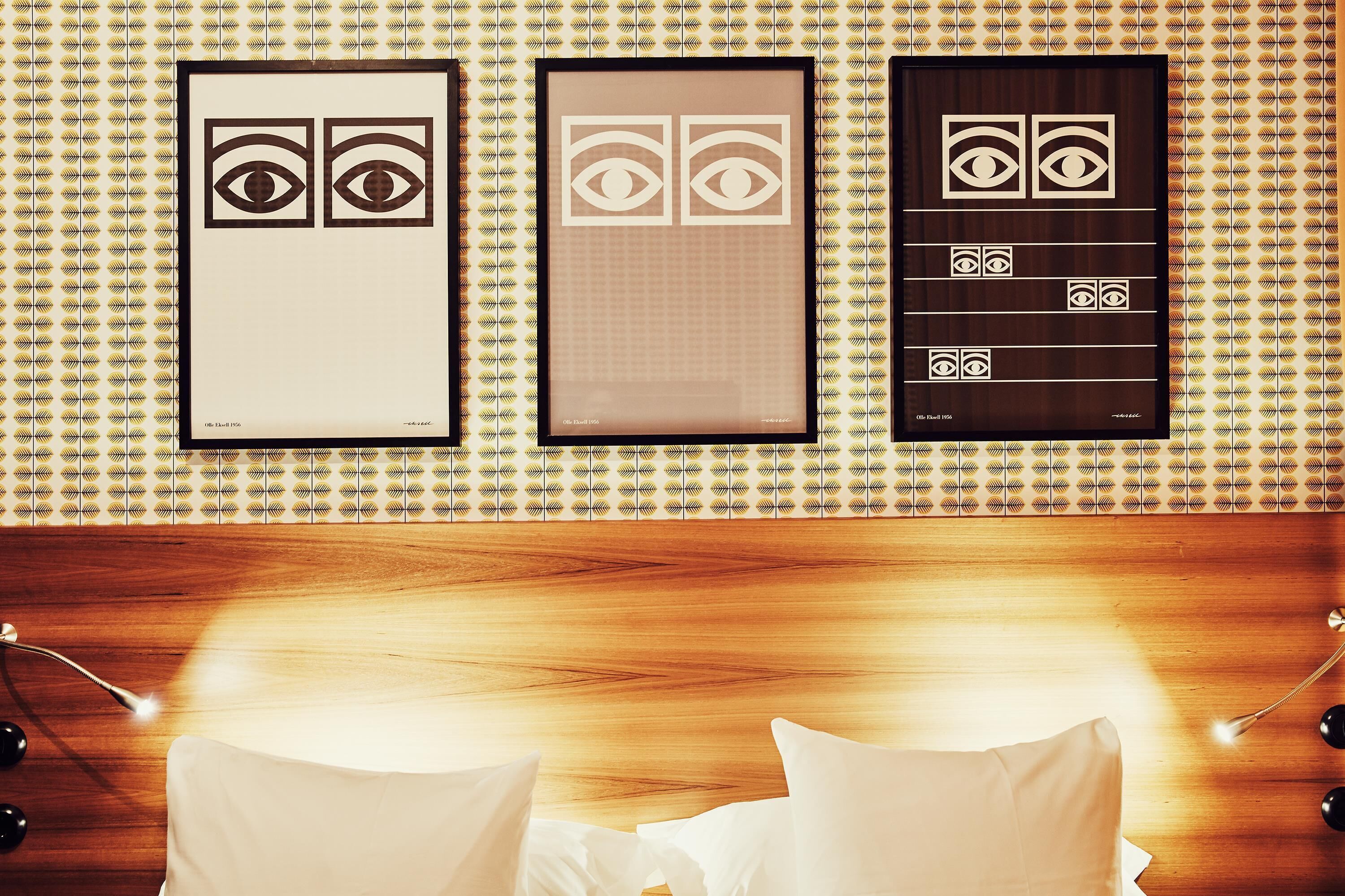 A hotel room with a patterned wallpaper, three framed artworks on the wall, and a neatly made bed with pillows.