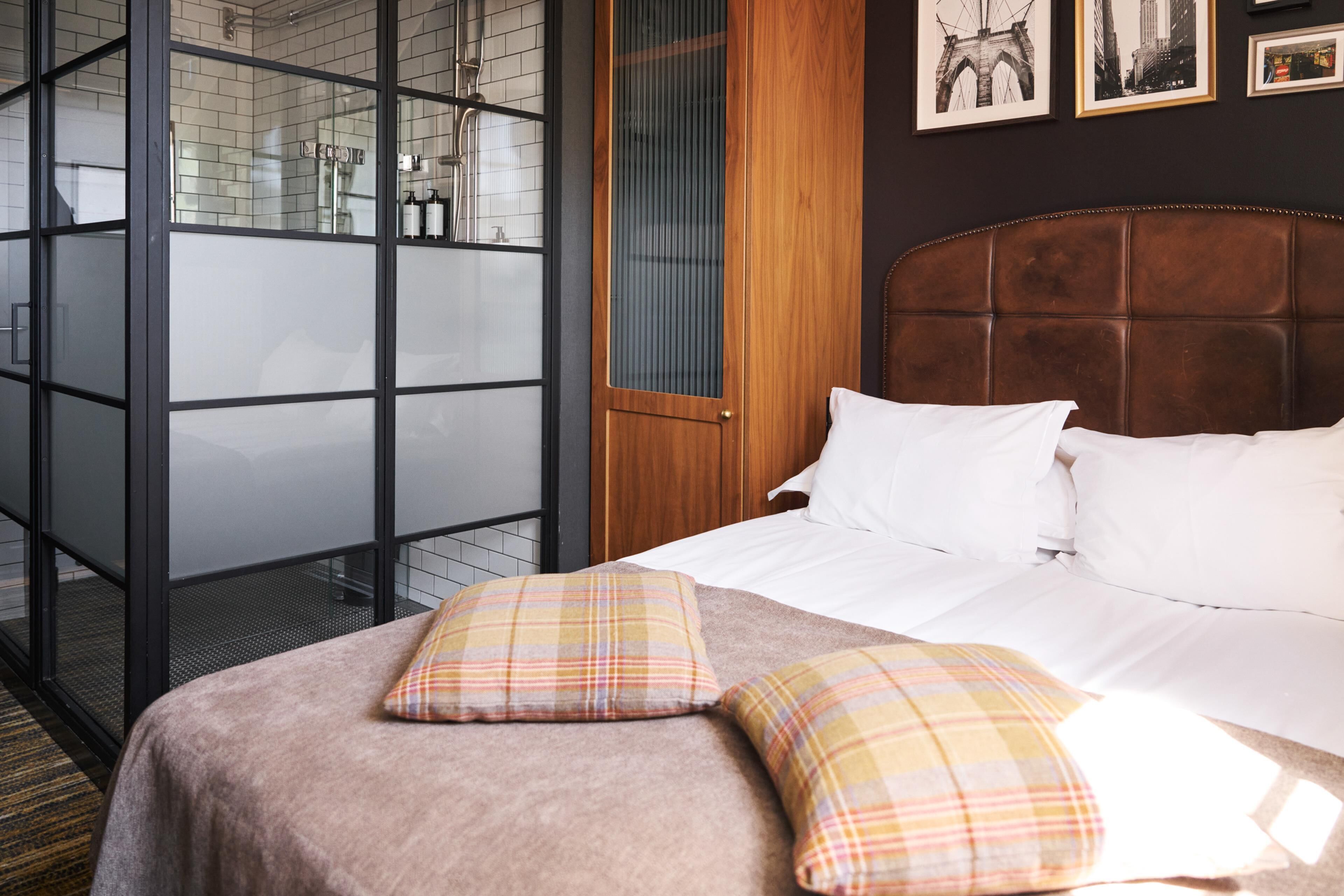 Quarto de hotel com uma cama, duas almofadas, cabeceira de madeira, divisória de vidro e uma porta.