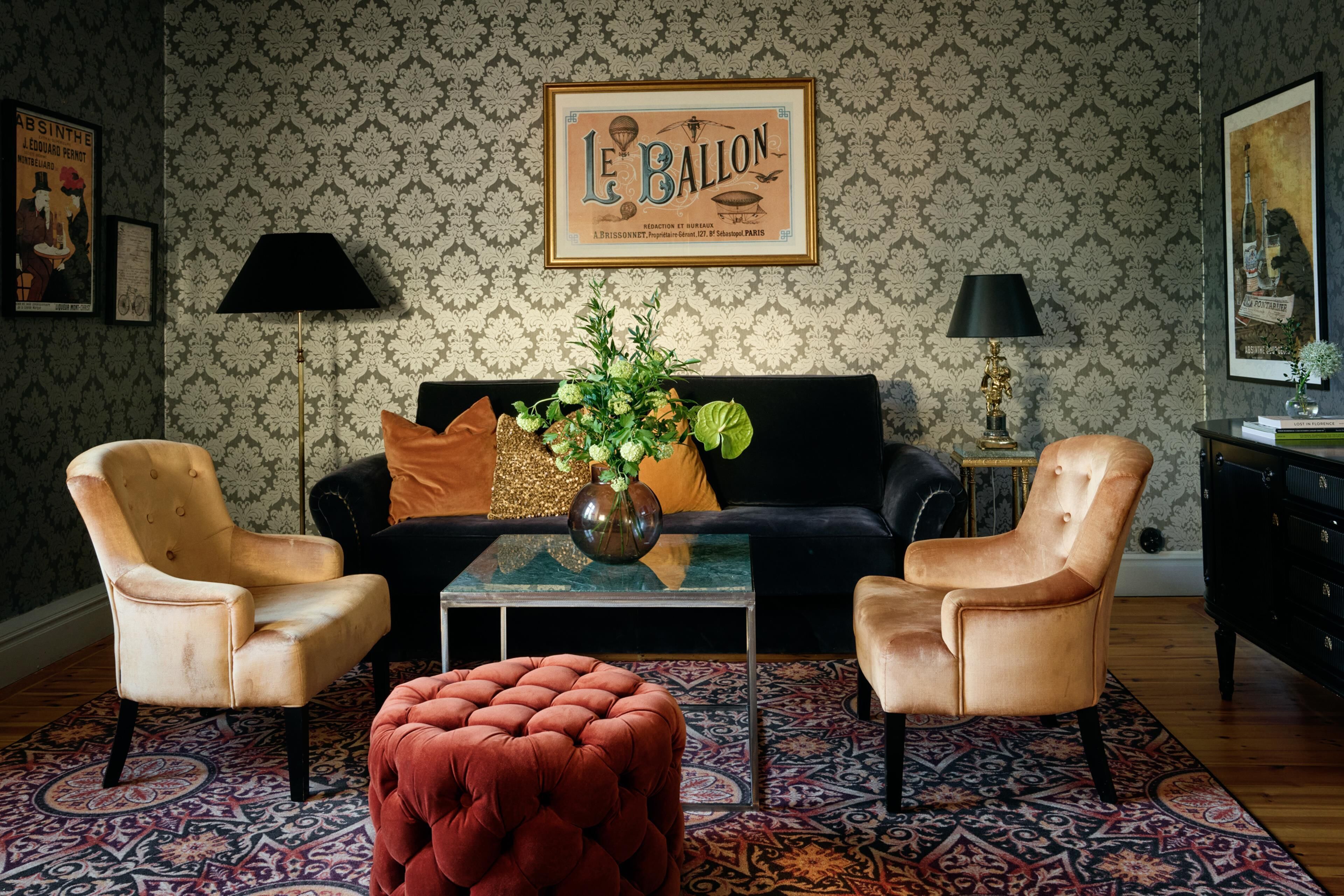 Ein Wohnzimmer mit schwarzem Sofa, zwei Sesseln, einem Couchtisch mit Vase, gemustertem Wandpapier und einem gerahmten Bild an der Wand.