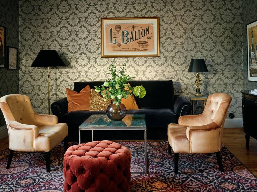 A living room with a black sofa, two armchairs, a coffee table with a vase, patterned wallpaper, and a framed picture on the wall.