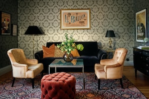 Uma sala de estar com um sofá preto, duas poltronas, uma mesa de centro com um vaso, papel de parede estampado e um quadro na parede.
