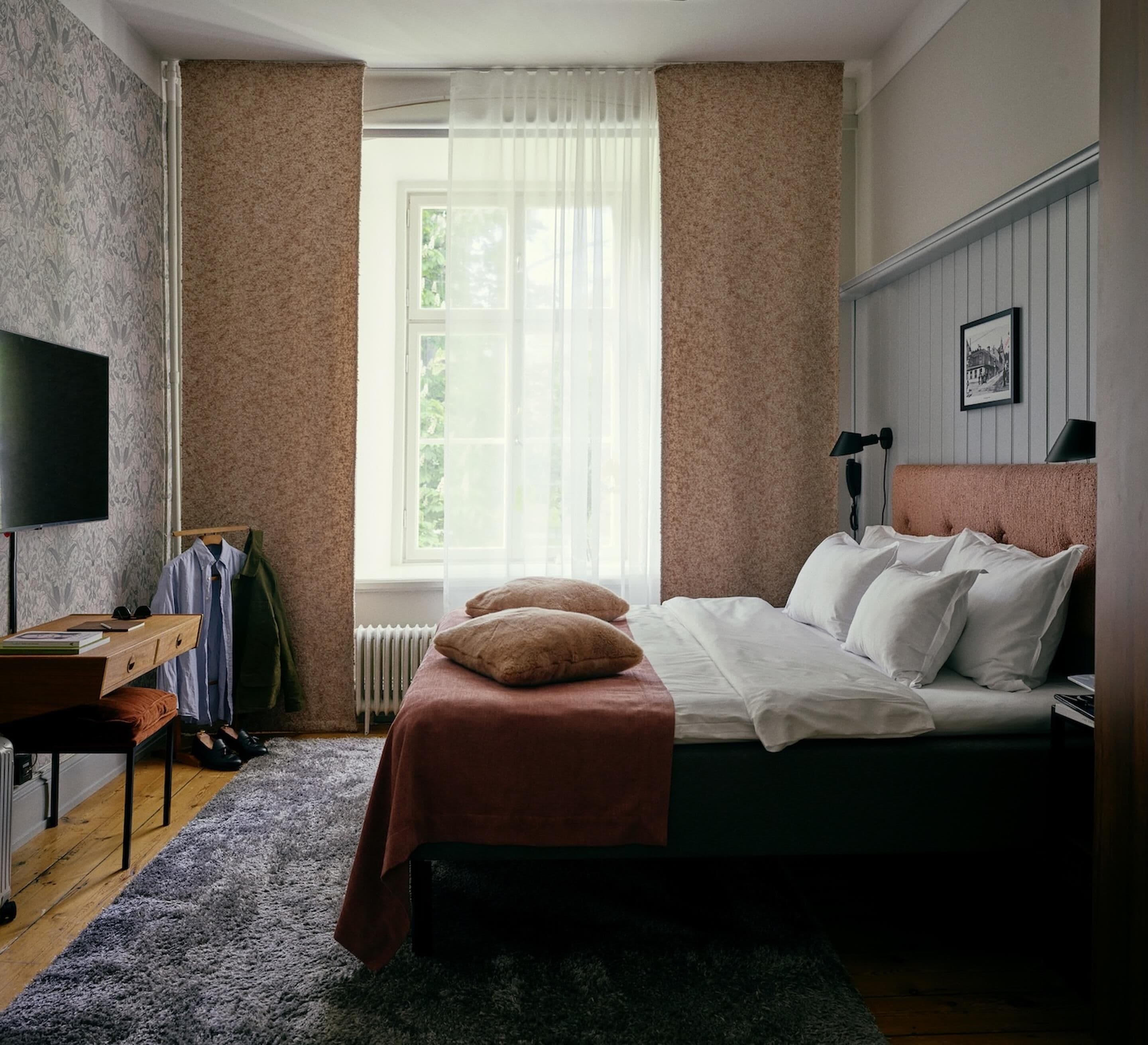 A bedroom with a bed, a desk, a chair, a TV, and a window with curtains.