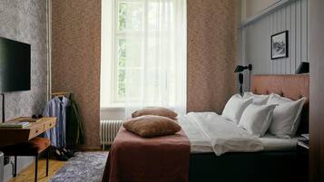 A bedroom with a bed, a desk, a chair, a TV, and a window with curtains.