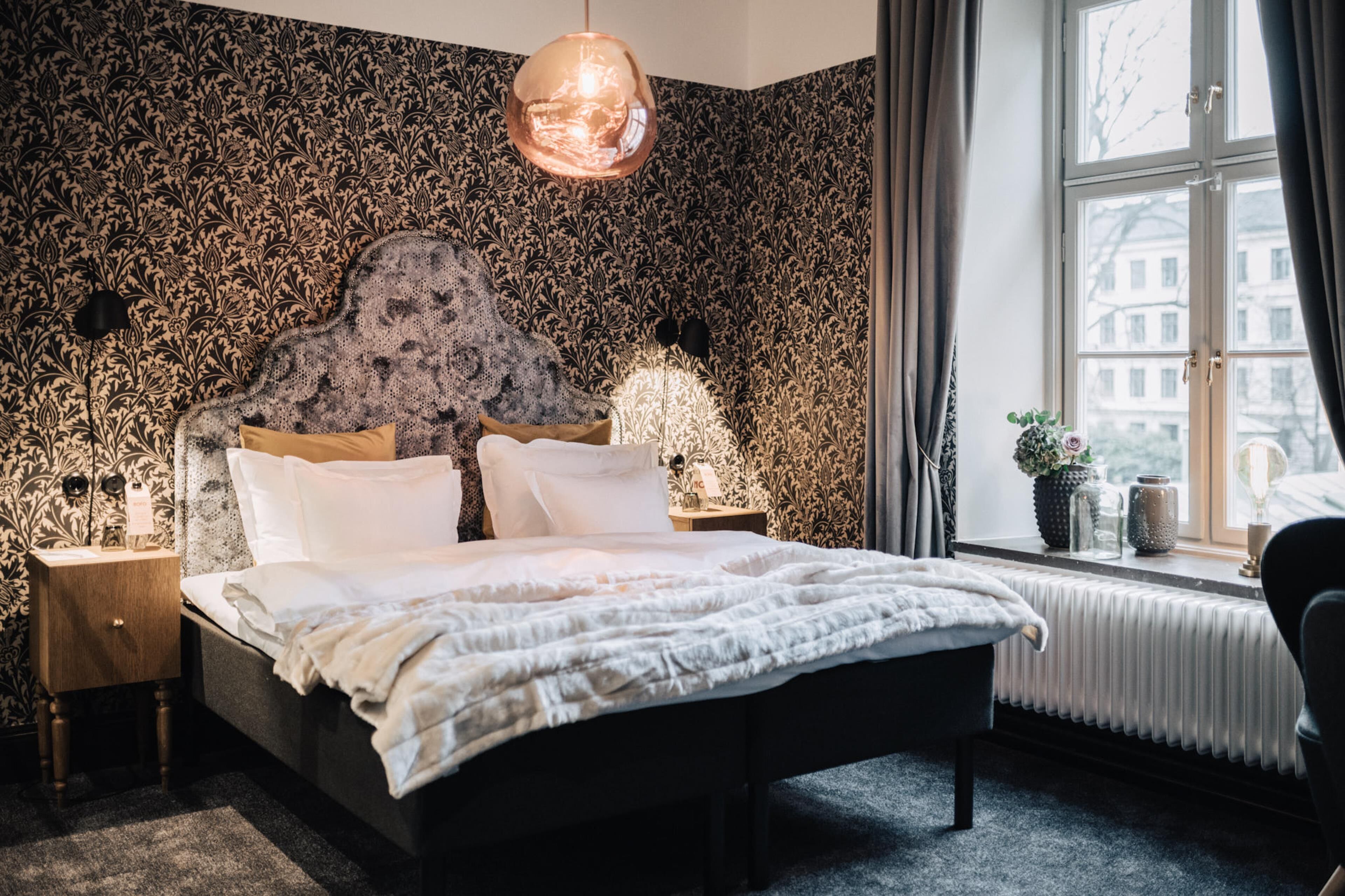A bedroom with a patterned wallpaper, a bed with white bedding, a hanging light fixture, and a window with a view of buildings.