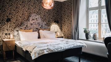 A bedroom with a patterned wallpaper, a bed with white bedding, a hanging light fixture, and a window with a view of buildings.