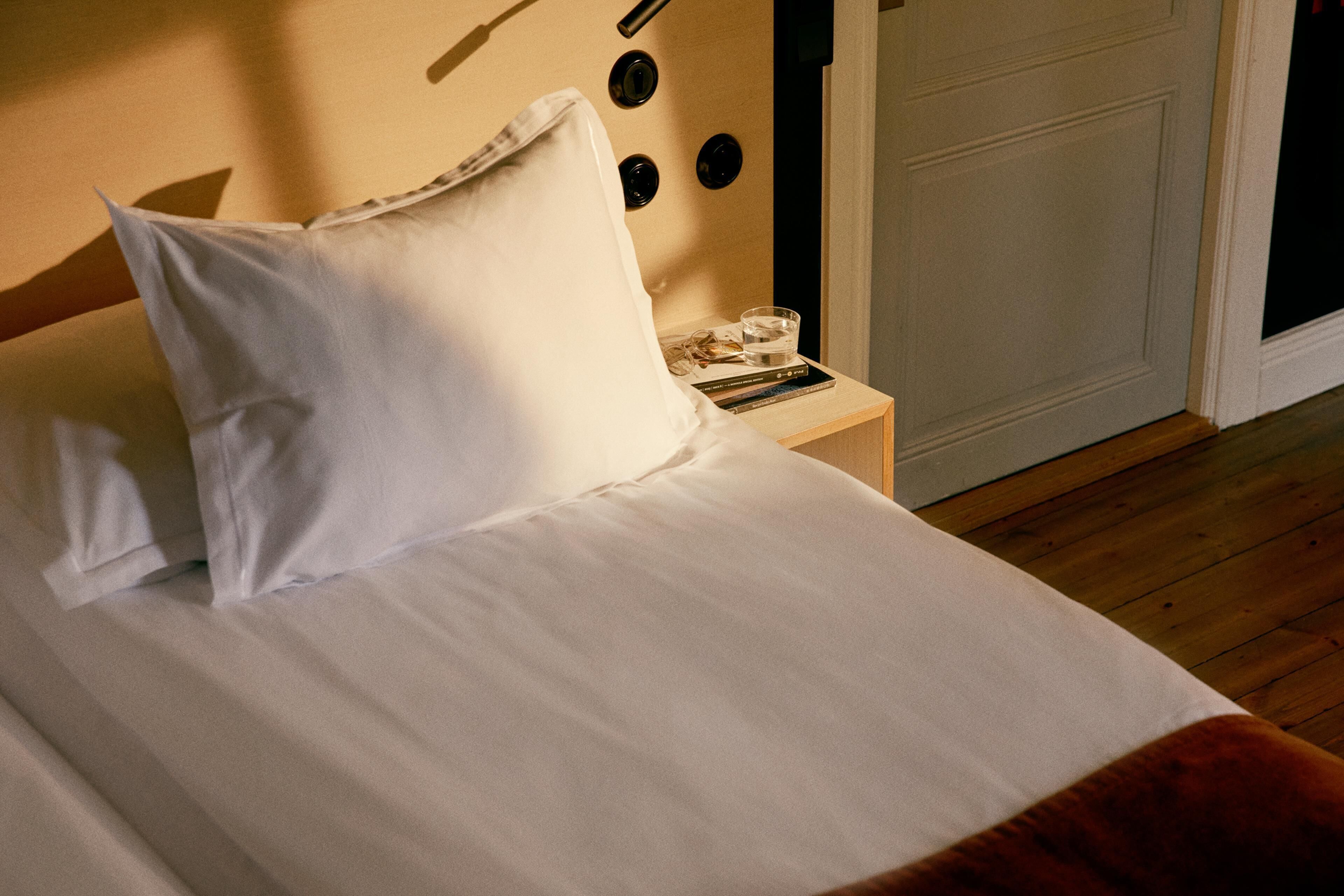 A neatly made bed with white linens and a pillow, a bedside table with a glass of water and books, and a wooden floor.