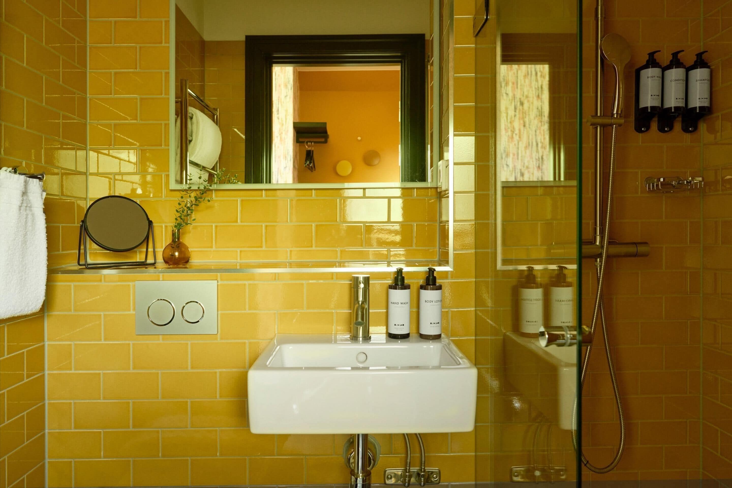 Baño con pared de azulejos amarillos, un lavamanos blanco, un espejo y artículos de tocador en la pared.