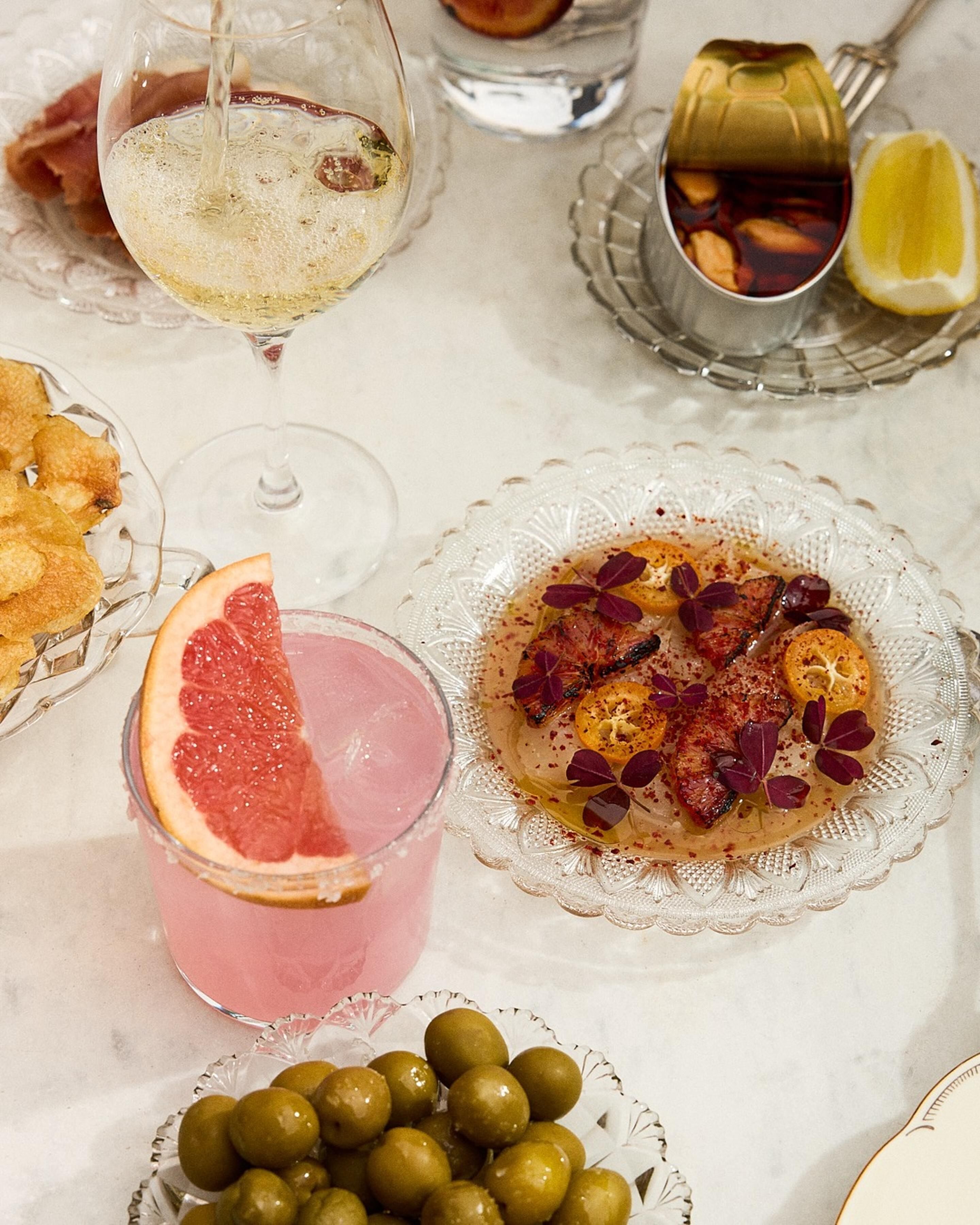 Una mesa con diferentes alimentos y bebidas, incluyendo una copa de vino blanco, una bebida rosada con rodaja de pomelo, un plato con carne y cítricos, y un cuenco de aceitunas.