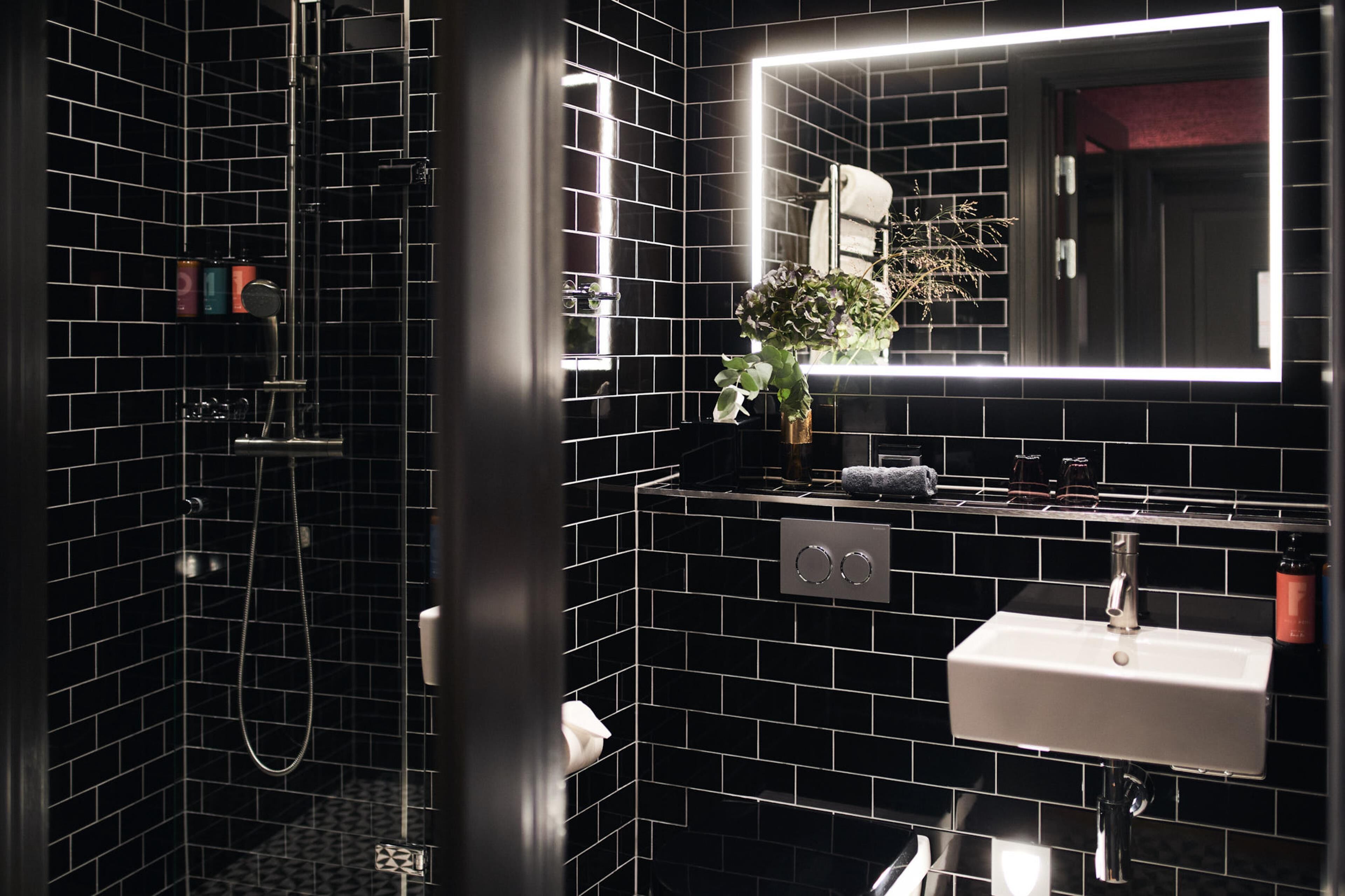 A modern bathroom with black tiled walls, a white sink, a handheld showerhead, and a mirror with a light.