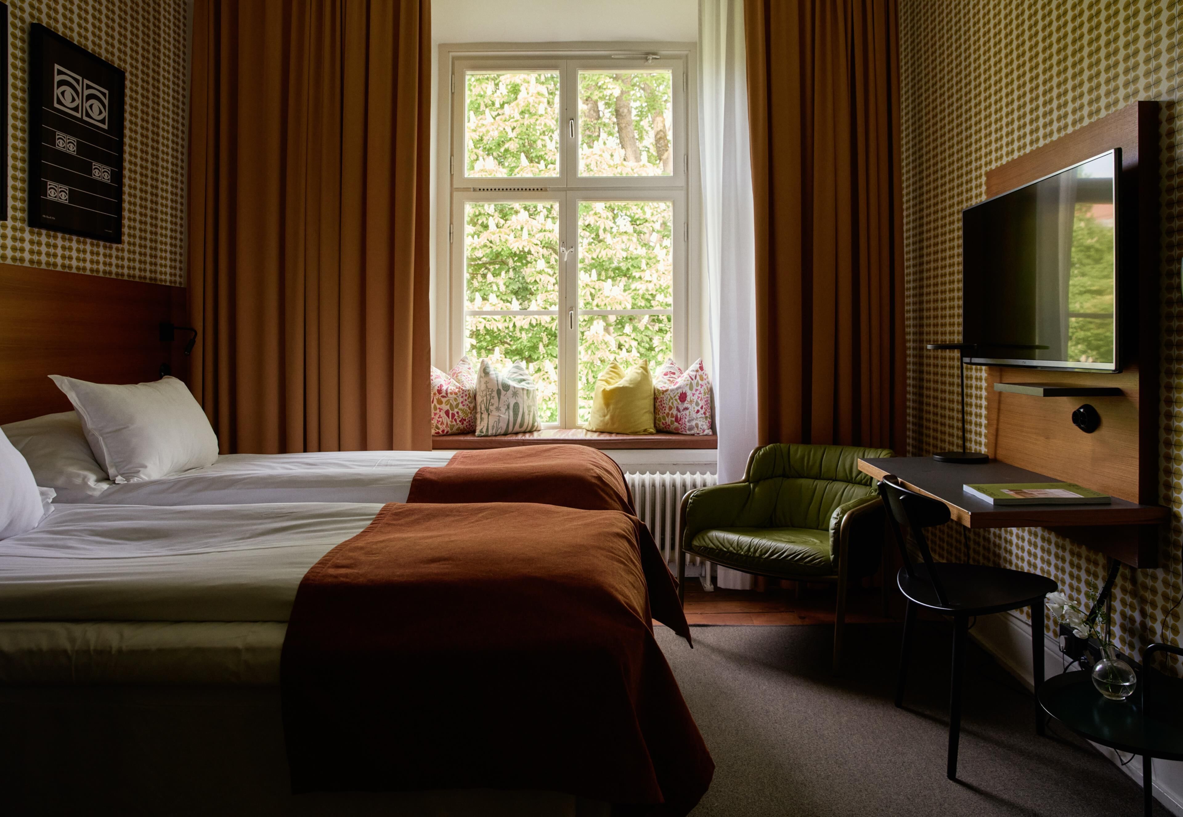 A hotel room with a bed, a desk with a chair, a television, and a window with a view of trees.